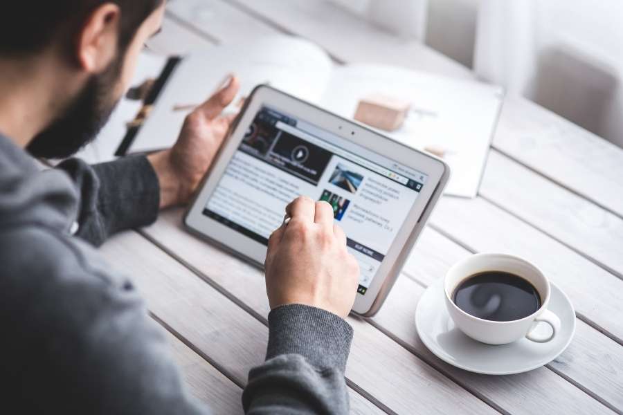 Un homme utilise une tablette tactile sur une table en bois lumineuse pour lire des articles et repérer une fausse information en ligne.